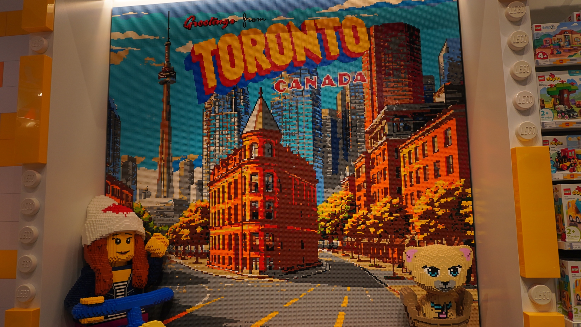 Large LEGO mosaic mural of the Toronto skyline with bold 'TORONTO CANADA' lettering, built from colorful bricks, flanked by display shelves on both sides.