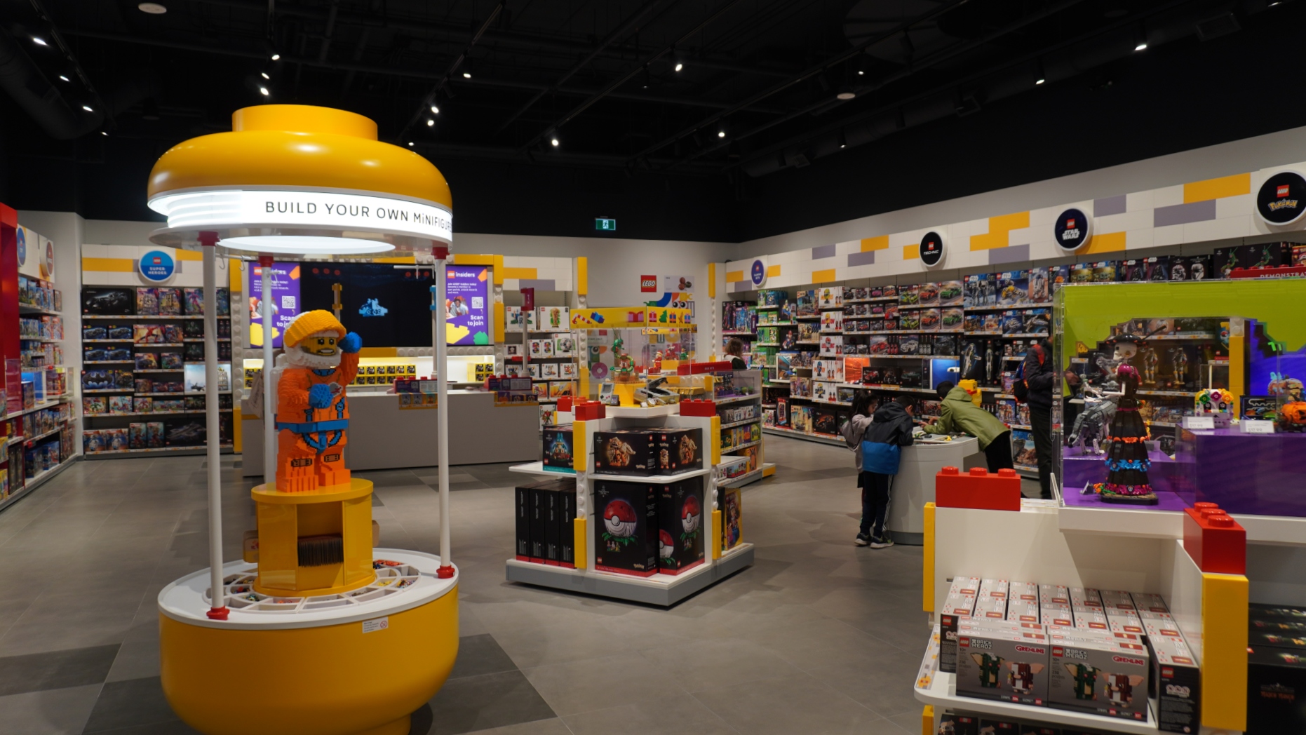 Bright LEGO store interior with a large yellow ‘build your own minifigure’ station in the foreground and shelves of sets along the walls, shoppers in the background.