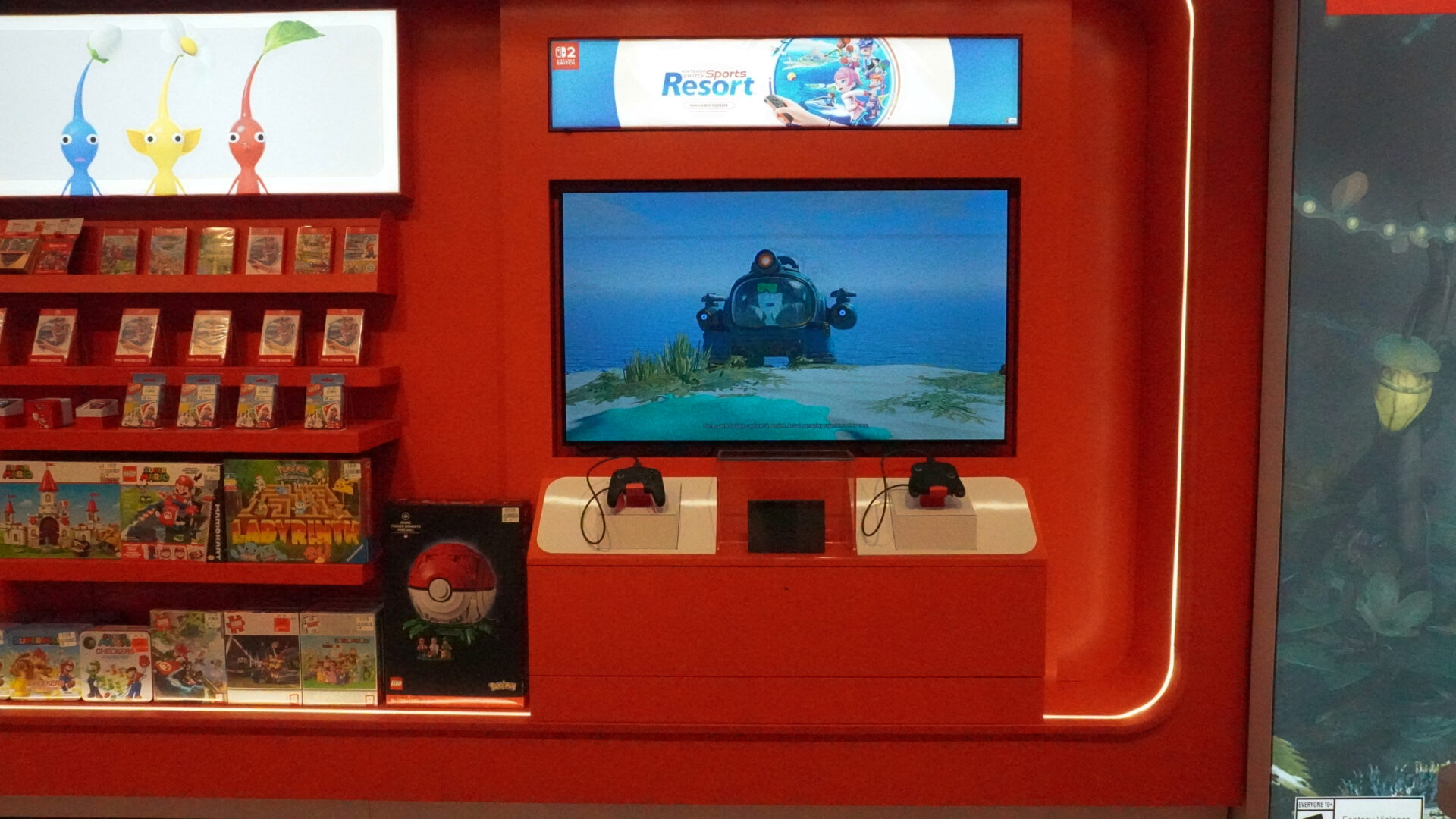 Nintendo-Store-3 | iPhone in Canada Red gaming kiosk with a screen showing an underwater submarine game; shelves of game cases on the left and two controllers on white mounts below the screen.