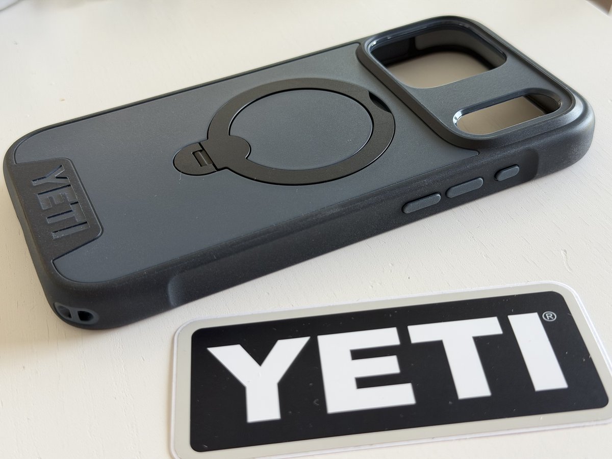 Empty navy YETI LoadOut iPhone case at an angle showing its side button covers, a bottom lanyard tie slot and the ring stand folded, next to a YETI sticker